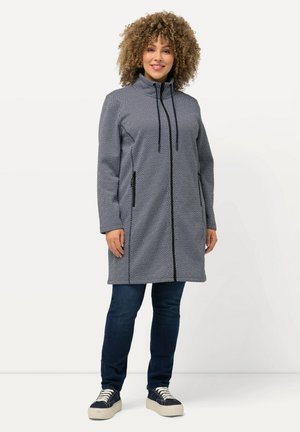 Gray and white patterned coat with a high collar, drawstring, front zippers, and side pockets, paired with dark jeans and sneakers.