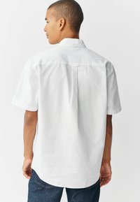 Camisa blanca de manga corta con cuello abotonado, que presenta un detalle de pliegue en la parte posterior. Fabricada en un tejido texturizado, con un corte de ajuste relajado.