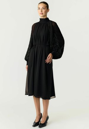 Femme portant une robe midi noire avec un col montant, des manches longues transparentes texturées et des talons noirs, debout devant un fond blanc.