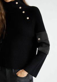 Maglione nero a maglia con collo alto, caratterizzato da una texture a costine, quattro bottoni metallici sulla spalla e dettagli in tessuto a contrasto sulle maniche.