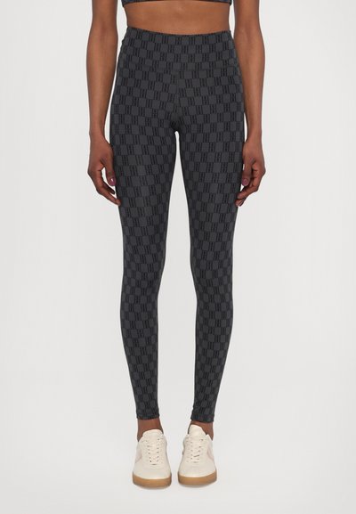 Hochtaillierte, schwarze Leggings mit einem ganzflächigen Musterdesign des Logos. Weiches, dehnbares Material mit einer glatten Textur. Kombiniert mit hellen Sneakern.