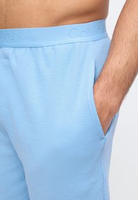 Κοντινό πλάνο ενός ατόμου που φοράει γαλάζια υφασμάτινα σορτς Calvin Klein, με το χέρι να στερεώνεται μέσα στην πλαϊνή τσέπη.