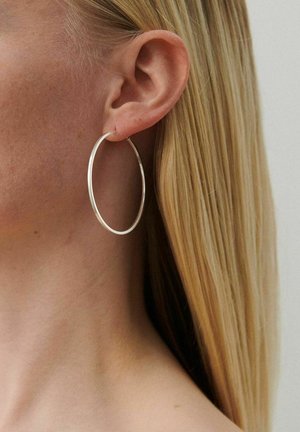 Zbliżenie na lewe ucho osoby z długimi blond włosami, noszącej duże, cienkie srebrne kolczyki-koła na neutralnym tle.