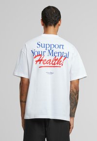 White cotton t-shirt featuring block text on the back in blue and red, reading "Support Your Mental Health!" with a logo below.