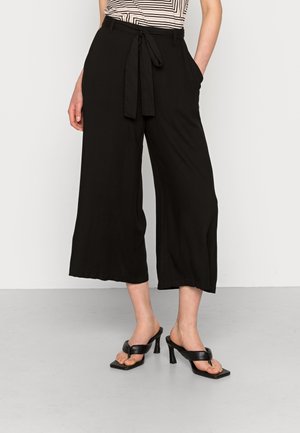 Femme portant un pantalon noir coupe large et courte avec ceinture à nouer, un haut rayé rentré, et des sandales noires à talons aiguilles, debout sur un sol gris.