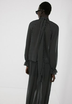 Persona con un vestido negro de manga larga con lunares, puños con volantes y un lazo en el cuello alto, de pie frente a un fondo claro y liso.