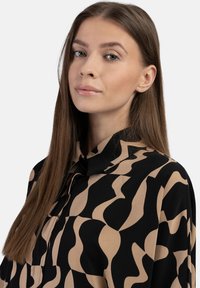 Schwarze Bluse mit einem beigen abstrakten wellenförmigen Muster, die einen Kragen hat und eine glatte Textur aufweist.