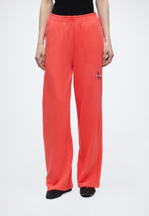 Person iført løse koralfarvede joggingbukser med elastisk talje og lille Calvin Klein-logo på venstre lår, kombineret med sorte sko og sort skjorte.
