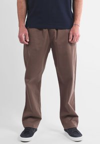 Pantalon marron léger à coupe décontractée, présentant une texture lisse et des plis devant, associé à des baskets en toile bleu marine.