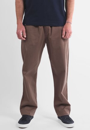 Pantalon marron léger à coupe décontractée, présentant une texture lisse et des plis devant, associé à des baskets en toile bleu marine.