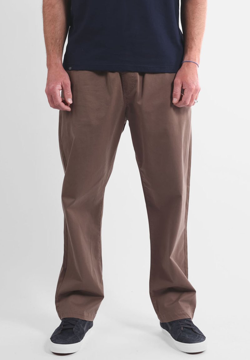 Pantalon marron léger à coupe décontractée, présentant une texture lisse et des plis devant, associé à des baskets en toile bleu marine.