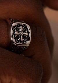 Zilveren ring met een ingewikkeld zwart ontwerp, met een vierkante bovenkant met een kruispatroon en opengewerkte details aan de zijden.