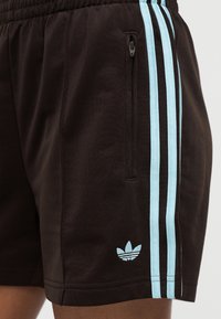 Κοντινό πλάνο μαύρων αθλητικών σορτς με τρεις ανοιχτόμπλε ρίγες, τσέπη με φερμουάρ και λογότυπο Adidas σε ανοιχτό μπλε κοντά στο τελείωμα.