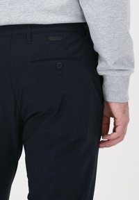 Navy trousers made of smooth fabric, featuring a back pocket with a button and a small brand tag above the pocket. Accompanied by a grey sweater.