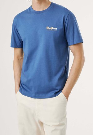 Homme portant un t-shirt bleu Pepe Jeans et un pantalon beige clair, les mains dans les poches, sur un fond uni.