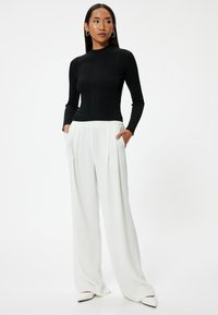 Schwarzes, geripptes Langarmoberteil kombiniert mit weißen, hoch taillierten, plissierten Hosen. Schmaler Schnitt, glatte Textur, dazu spitz zulaufende weiße Schuhe.