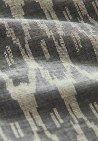 Textiel met een lineair patroon van witte figuren op een getextureerde donkergrijze achtergrond, waarbij unieke vormen en subtiele variaties in de weving worden benadrukt.