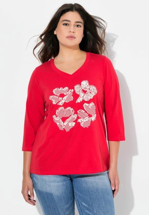 T-shirt en coton rouge avec un col en V et des manches trois-quarts, présentant un imprimé graphique floral et à rayures blanches sur le devant.