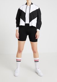 Svart och vit skuren jacka med geometrisk design över en vit t-shirt, matchad med svarta shorts och vita strumpor med röda ränder.