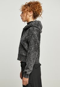 Sudadera negra recortada en un tejido desgastado y texturizado, con capucha ajustable y puños acanalados. Expuesta de lado sobre un fondo neutro.
