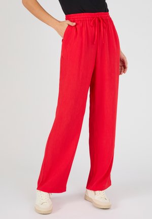 Person trägt lockere rote Hose mit elastischem Bund und Kordelzug, kombiniert mit weißen Turnschuhen, steht vor einfachem Hintergrund.