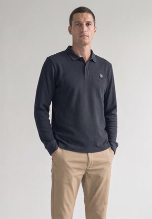 Homme debout, les mains dans les poches, portant un polo à manches longues sombre et un pantalon beige sur un fond clair uni.