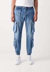 Pantalones cargo en denim azul medio, con múltiples bolsillos laterales, puños ajustados y un ajuste relajado para facilitar el movimiento.