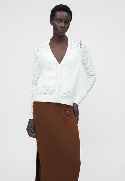 Γυναίκα που φοράει λευκή πουλόβερ με κουμπιά και σχέδια με τρύπες και καφέ φούστα ψηλόμεση με πλαϊνή σχισμή και λεπτομέρειες με κορδόνια.