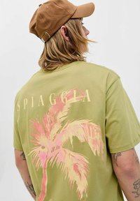 Oliven grøn t-shirt med lyserødt palmetræ-tryk på ryggen. Har rund halsudskæring og afslappet pasform, kombineret med en brun kasket.
