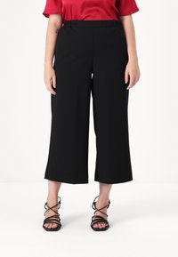 Siledest mustad culotted, millel on lai jalgeosa lõige ja elastne vöökoht, koos mustade sandaalidega õhukeste rihmadega. Näha on punane topp.