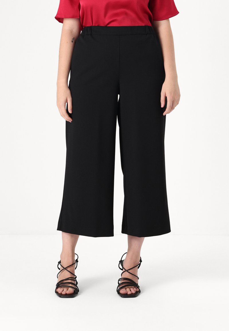 Siledest mustad culotted, millel on lai jalgeosa lõige ja elastne vöökoht, koos mustade sandaalidega õhukeste rihmadega. Näha on punane topp.