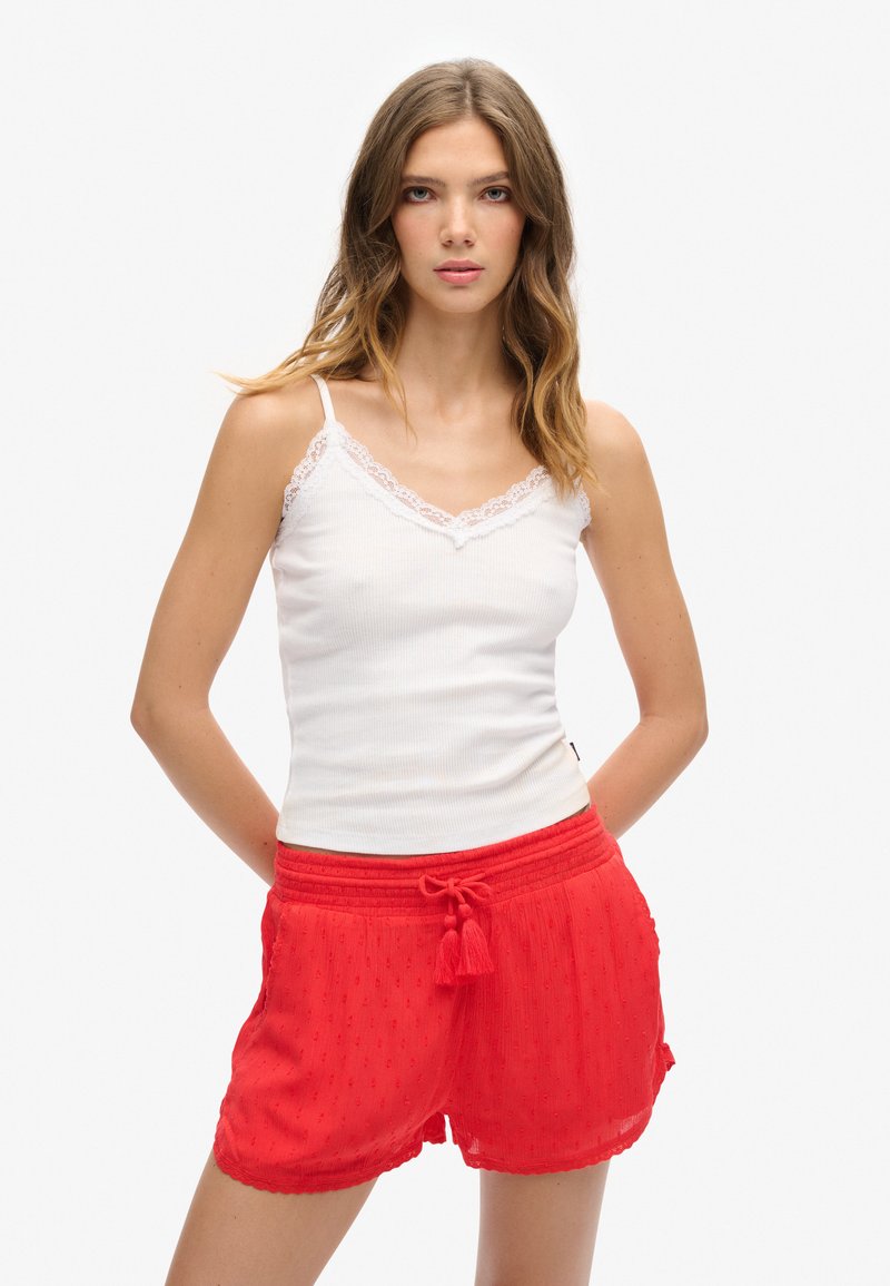 Top de tirantes blanco con acento de encaje, combinado con pantalones cortos rojos texturizados que tienen una cintura fruncida y un detalle de flecos atado.