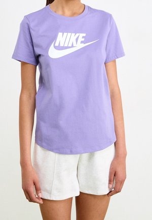 Personne portant un t-shirt Nike violet avec un logo blanc et un short gris clair, debout devant un fond blanc uni.