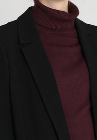 Žena v burgundském pleteném roláku pod černým sakem, které je zkrácené u krku a ramen.