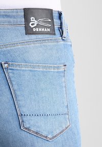 Ljust blå denimjeans med en triangulär bakficka och en svart läderetikett med vita saxar och varumärkesnamnet "DENHAM."