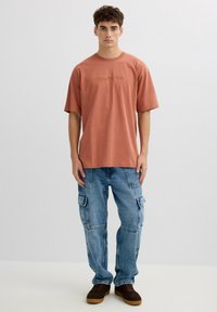 Joven de pie con una camiseta de gran tamaño color óxido, jeans cargo azul claro con bolsillos y zapatillas marrón oscuro, sobre un fondo liso.