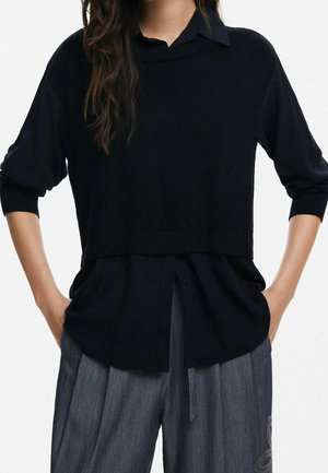 Femme portant un chemisier noir à manches longues superposé avec les manches retroussées et un pantalon ample gris brodé d'un motif floral.