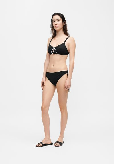 Γυναίκα που στέκεται φορώντας μαύρο μπικίνι με λευκές λεπτομέρειες φιόγκων και μαύρα σανδάλια σε μονόχρωμο λευκό φόντο.