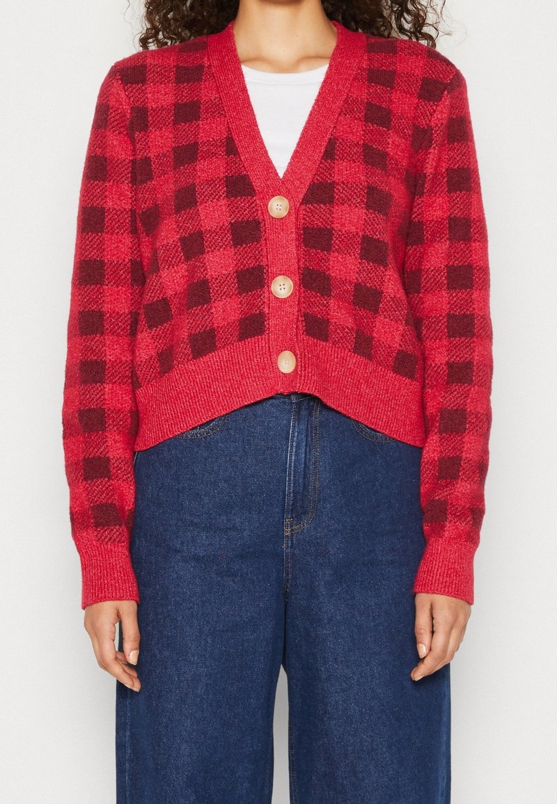 Punainen ja tumma ruudullinen neulecardigan, cropped-malli, V-pääntie, kolme nappia edessä, ribattu hihansuut ja helma. Yhdistetty tummansinisiin leveälahkeisiin farkkuihin.