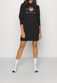 Robe à capuche noire avec une poche avant, ornée d'un design de logo coloré. Le matériau semble doux ; longueur genou avec des manches longues.