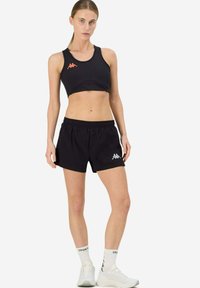 Completo sportivo nero composto da reggiseno e pantaloni corti realizzati in tessuto leggero. Presenta il logo Kappa in arancione e grigio. Design semplice con vita elasticizzata.