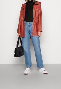 Rode jas van nep leer met capuchon, zwarte coltrui, lichtblauwe jeans met rechte pijpen, zwarte handtas en witte sneakers met rode accenten.