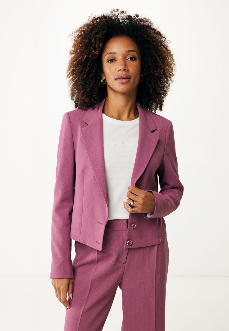 Blazer rose ajusté avec des épaules structurées, fermeture à deux boutons et tissu texturé. porté sur une chemise blanche avec un logo sur le devant.