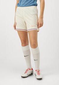 Nike krēmkrāsas futbols šorti ar rozā logotipu, ziliem un sarkaniem svītrojumiem gar apakšmalu, saskaņoti ar baltām ceļgalu augstām zeķēm un futbolu zābakiem.