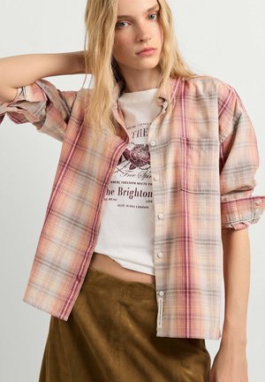 Jonge vrouw met lang blond haar die een beige en roze geruite blouse draagt over een wit grafisch T-shirt en een olijfgroene bruine rok.