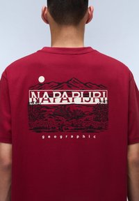 Camiseta de algodón roja con un gráfico de paisaje montañoso en negro y el texto "NAPAPIJRI" y "geográfico" debajo, en la parte posterior.