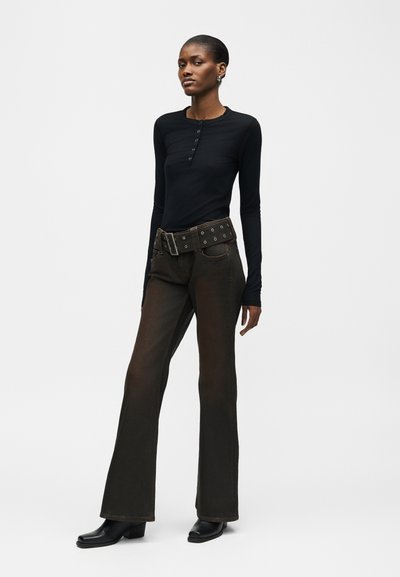 Γυναίκα όρθια, φοράει εφαρμοστό μαύρο μακρυμάνικο τοπ, σκούρο καμπάνες παντελόνι με φαρδιά ζώνη και μαύρα μποτάκια αστραγάλου, μπροστά σε απλό φόντο.