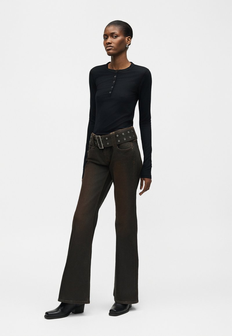 Woman standing, wearing a fitted black long-sleeve top, dark flared pants with a wide belt, and black ankle boots, against a plain background.