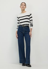 Frau, die vor einem schlichten weißen Hintergrund steht und einen weißen Pullover mit schwarzen Streifen, hoch geschnittene blaue Jeans, einen schwarzen Gürtel und schwarze Schuhe trägt.