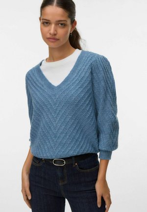 Lichtblauwe gebreide trui met een V-hals en een textuurpatroon in chevronstijl, gecombineerd met een wit shirt, gedragen met een donkere denim jeans en een zwarte riem.